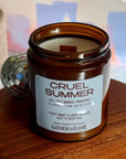 Candle jar labeled 'Cruel Summer' on a wooden surface with a blurred background