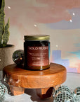 Candle labeled 'Gold Rush' on a wooden stand with decorative items and a plant in the background.