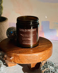 Candle jar labeled 'Gold Rush' on a wooden stand with decorative items and a colorful background