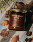 Candle labeled 'Beachside Fire' on a surface with shells and driftwood