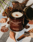 Candle labeled 'Beachside Fire' on a striped fabric with shells and wood, evoking a beachside theme.