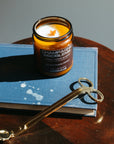 Candle labeled 'Champagne Problem' on a book with gold wick trimmers on a wooden surface