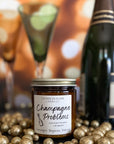 A jar candle with a label reading 'Champagne Problems' surrounded by gold metallic balls, a bottle of champagne, and champagne flutes.