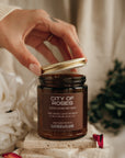 Hand opening a candle jar labeled 'City of Roses' with decorative elements in the background.
