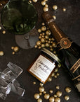A jar candle with a label reading 'Champagne Problems' surrounded by gold metallic balls , broken glass, champagne bottle and champagne flutes