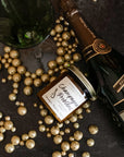 A jar candle with a label reading 'Champagne Problems' surrounded by gold metallic balls, champagne bottle and champagne flutes