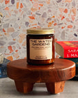 Candle labeled 'The Water Garden' on a wooden stand with books and a bust statue in the background.
