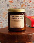 Candle jar labeled 'The Water Gardens' by 'Gather in Flame' on a wooden stool with a patterned wall in the background.