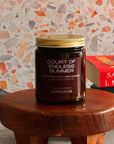 Candle jar labeled 'Court of Endless Summer' on a wooden stool with a colorful wall and books in the background