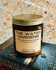 Candle labeled 'The Water Gardens' on top of books with a patterned wall background