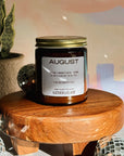 Candle jar labeled 'August' on a wooden stool with a blurred background