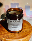 Candle in an amber jar labeled 'August' on a wooden stand with a blurred background.