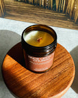 Candle in a glass jar labeled 'Once Upon a Churro' on a wooden coaster with a reflective surface in the background.