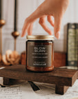 Jar of 'Slow Burn' candle on a wooden stand with a hand reaching towards it, surrounded by books and candles.