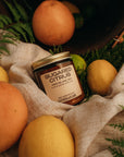 Jar of 'Sugared Citrus' candle surrounded by fruits and foliage on a textured fabric background