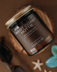 Candle jar labeled 'West Coast Hottie' by Gathered Flame on a brown background with starfish and a flower.