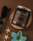 Jar of 'West Coast Hottie' candle on a wooden coaster with decorative items on a brown background