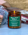 Candle in a amber jar with a green label on a surface with books and a blanket.