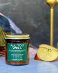 Jar of 'All Too Well' candle with a green label on a textured surface with apples and decorative elements.
