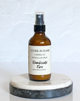 A brown amber spray bottle labeled 'Gather In Flame Candle Co. Beachside Fire Room & Car Spray' placed on a white marble surface.