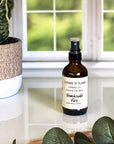 A brown amber spray bottle labeled 'Beachside Fire' by Gather In Flame Candle Co. on a white marble surface with a window and plant in the background.