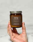 Amber glass jar candle labeled Book Club held in a hand with a white background.