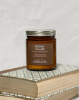 Amber glass jar candle labeled Book Club on a stack of books with a white background.