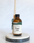 A brown glass reed diffuser bottle with a label reading "Bougie Hotel", placed on a white marble round tray, with reeds inserted into the bottle for diffusing the fragrance.