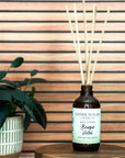 A brown glass reed diffuser bottle with a label reading "Bougie Hotel", placed on a wood surface, with plant and wood slat background.