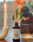 A brown glass reed diffuser bottle with a label reading "Bougie Hotel", placed on table with candle and flowers in a vase in the background.