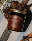 Jar of 'Campfire Marshmallows' candle on a striped surface with wooden elements and a plant.