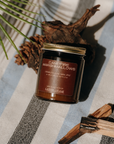 Jar of 'Campfire Marshmallows' candle on a striped fabric background with natural elements.