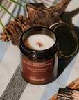 Campfire Marshmallows candle on a striped surface with natural elements.