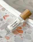 A perfume oil roller with a clear glass body and bamboo cap, labeled 'Champagne Problems', resting on a terrazzo surface.