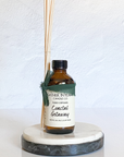 A Reed diffuser with amber glass bottle placed on a round white marble coaster. The label on the bottle reads 'Coastal Getaway'.