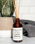 A brown glass reed diffuser bottle with a label reading "Coastal Getaway", placed grey surface, surrounded by plant and towels with white tile background.