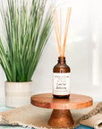 A brown glass reed diffuser bottle with a label reading "Coastal Getaway", placed on wood surface, with plant in the background.