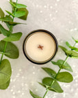 Overhead view of candle surface with wood wick surrounded by green leaves on a textured white background