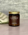 Jar of 'Cozy Cashmere' candle with a gold lid on a neutral background