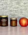 Two candles of different sizes labeled 'Cozy Cashmere' next to an apple on a textured surface.
