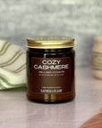 Jar of 'Cozy Cashmere' candle with gold lid on a surface with dried pampas grass, a soft blanket and mug in the background