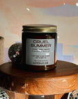 Candle labeled 'Cruel Summer' on a wooden stool with decorative items in the background