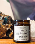 Amber jar candle labeled Don't Let the Hard Days Win on a wood surface with art in the background and large crystals.