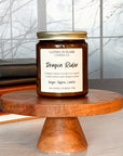 A jar candle labeled 'Dragon Rider' from Gather in Flame Candle Co.,' placed on a wooden pedestal, with books and a window in the background.