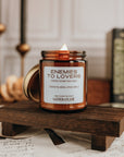 Candle labeled 'Enemies to Lovers' on a wooden stand with books and candles in the background.
