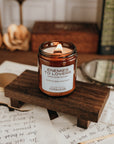 Candle in a jar labeled 'Enemies to Lovers' on a wooden stand with papers and a mirror in the background.