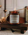 Candle labeled 'Enemies to Lovers' being lit on a wooden stand with books and candles in the background.