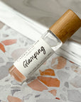 A perfume oil roller with a clear glass bottle and a bamboo cap, labeled 'Glamping', resting on a terrazzo surface.