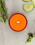 Lit candle topdown view on a light gray surface with green herbs and cucumber slices.