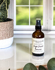 A brown amber spray bottle labeled 'High Desert Summer' by Gather In Flame Candle Co. on a white marble surface with a window and plant in the background.